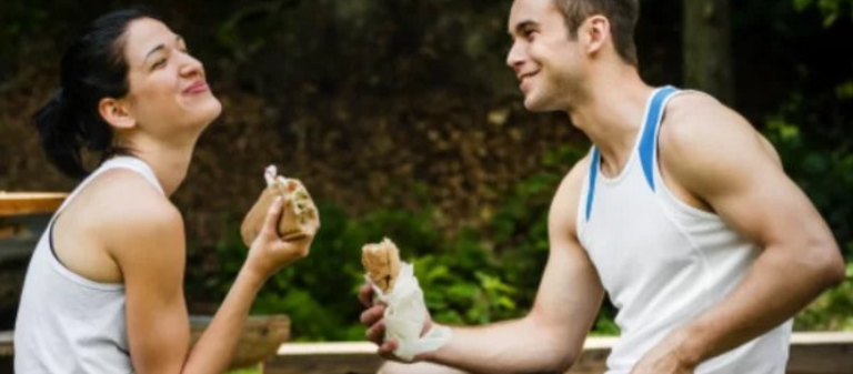 Man and woman in sports clothes sitting together, laughing and eating lunch – representing balance, joy, and the role of self-reflection in health and wellbeing