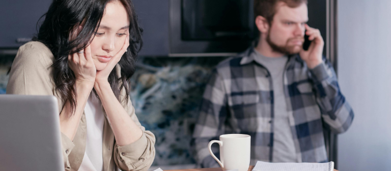 Woman looking at laptop with papers and bills scattered on the table, while man in the background speaks on the phone looking stressed, representing the pressures of being stuck in a stress response